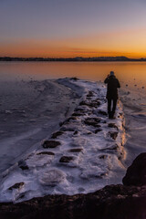 Silhouette of a Photographer Capturing the Golden Sunset Over a Frozen Lake. A person stands on icy shores, capturing the serene sunset over a tranquil, frozen lake.