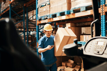 Young male warehouse worker using smartphone and carrying a box