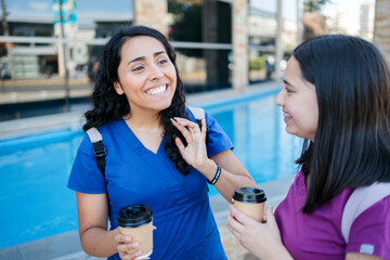 Two young hardworking latin nurses talking with cup of coffee after work. Female Healthcare workers leaving hospital