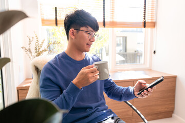 Asian man in casual clothes, holding coffee cup and using smartphone to checking email and social...