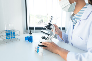 Scientist woman in medical face mask using microscope and test tube with blue liquid while working