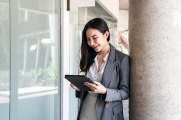 Fototapeta premium Asian businesswoman in suit, searching business data and reading text message on screen of digital tablet while standing