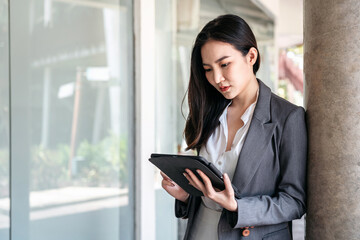 Asian businesswoman in suit, searching business data and reading text message on screen of digital tablet while standing