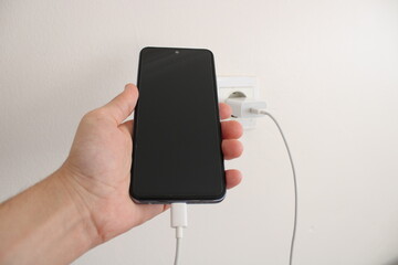Hand holding a charging smartphone. Electrical outlet in the background. fast charging and...