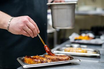 In a bustling professional kitchen, a chef meticulously applies glossy sauce to a grilled steak,...