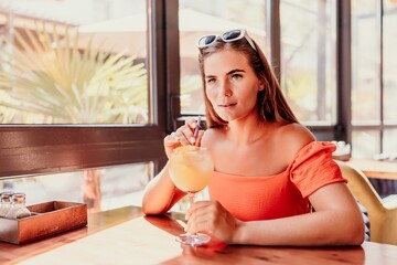 woman in a cafe seated, holding beverage. Dressed in an orange dress with her hair down. Bright...