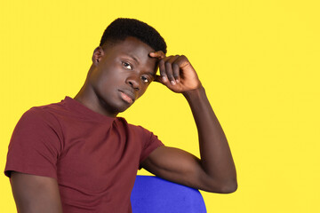 Young black man sitting on a blue chair wearing red t-shirt and yellow background, looking seriously at the camera with copy space. Thinking concept.
