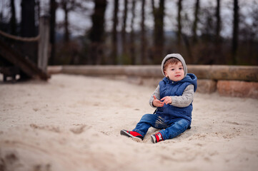 A young child dressed warmly in a vest and hat sits thoughtfully in the sand of a playground, capturing a moment of childhood tranquility and exploration