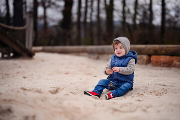 A young child dressed warmly in a vest and hat sits thoughtfully in the sand of a playground, capturing a moment of childhood tranquility and exploration
