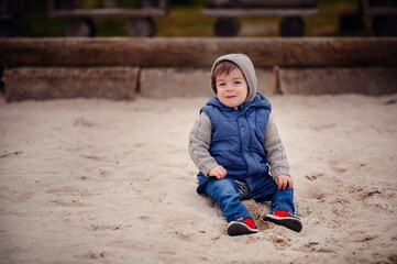 A young child dressed warmly in a vest and hat sits thoughtfully in the sand of a playground,...