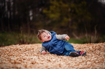Naklejka na ściany i meble A little boy lies in a bed of wood chips, his joy and playfulness captured in a natural outdoor setting, with a backdrop of trees