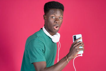 Young black man looking surprised with black headphones plugged into a mobile phone on a red background, with copy space.