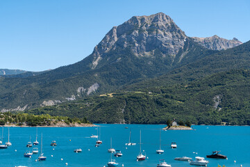 Lake of Serre-Poncon with the Chapelle Saint-Michel surrounded by boats during summer, Hautes-Alpes, France - obrazy, fototapety, plakaty