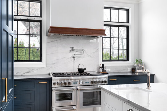 A luxurious kitchen detail with blue cabinets, a stainless steel stove, granite backsplash and countertop, and black framed windows. No brands or labels.