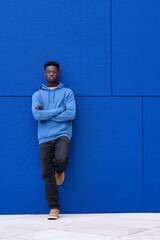 Attractive young black man leaning on a wall wearing blue sweatshirt, on a blue background, with copy space. Relaxation concept.