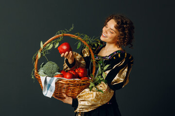 Portrait of a young adult woman dressed in a medieval dress holding a basket with vegetables and fruits. Harvest and healthy food concept. - 752557489