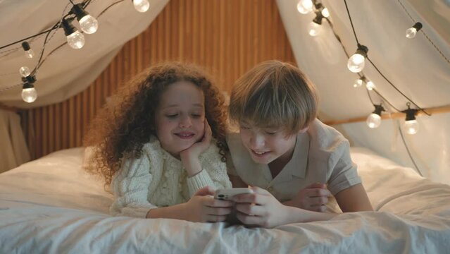 Cute sweet boy and girl lying in a tent with lights using smartphone telephone, watching photos or pictures, chatting laughing. Caucasian  siblings playing game on mobile phone at night, no sleeping