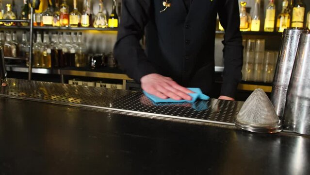 Barman in black waistcoat disinfects bar counter in pub. Bar equipment stands on surface with various alcohol in background