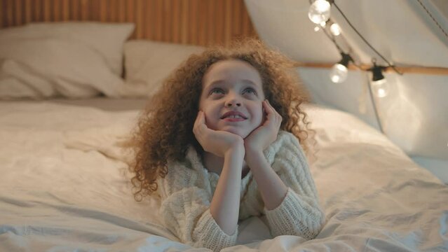 Attractive curly haired hispanic latin American ten-year-old girl lying on floor in play tent decorated with fairy lights dreaming thinking resting relaxing in modern bedroom at night, leisure 