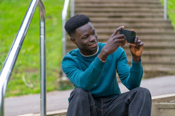 Attractive young black man with a blue t-shirt, smiling at the camera of his phone, taking a selfie sitting on some stairs in a park in the city of Bilbao. Happiness concept.