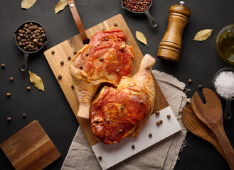 Raw chicken drumsticks seasoned on a wooden board, accompanied by salt and peppercorns, viewed from...