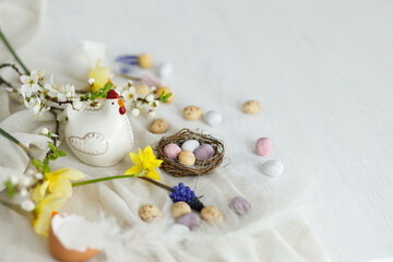 Happy Easter! Stylish easter chocolate eggs in nest, spring flowers, chicken figurine on white rustic wooden table. Easter modern simple banner, space for text. Seasons greetings