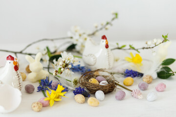 Fototapeta na wymiar Happy Easter! Stylish easter chocolate eggs in nest, spring flowers, chicken figurine on white rustic wooden table. Easter modern simple banner, space for text. Seasons greetings