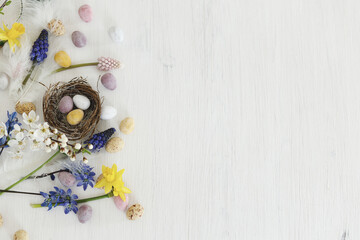 Easter rustic flat lay. Colorful easter chocolate eggs in nest, spring flowers, feathers border composition on white wooden table. Space for text. Happy Easter! Seasons greetings
