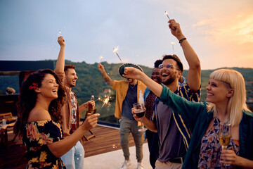 Cheerful friends using sparklers while dancing during summer party on patio at sunset.