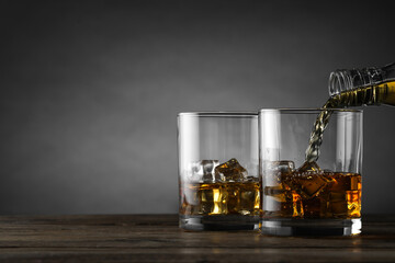 Pouring whiskey into glass with ice cubes at wooden table against grey background, space for text