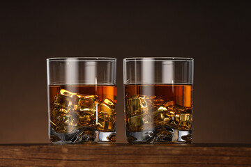 Whiskey with ice cubes in glasses on wooden table against brown background, closeup