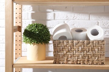 Toilet paper rolls in wicker basket and floral decor on wooden shelf against white brick wall