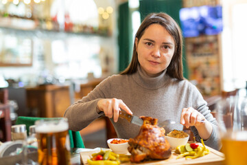 Positive brunette having lunch in Viennese cafe with traditional dish Schweinsstelze, crispy roasted ham hock with vegetable side dishes and glass of beer. Local cuisine - obrazy, fototapety, plakaty