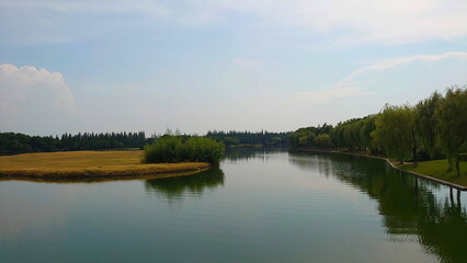 Zhujiajiao Ancient town, Shanghai, China - July 25, 2019: peaceful lake surrounded by willow trees and grassland with a wooden bridge in the background under a bright blue sky