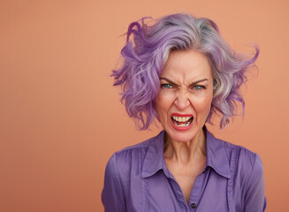 Portrait of an angry woman 60 years old with purple hair in purple clothes on an isolated peach color background. Aggressive woman grandmother