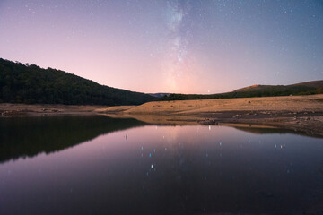 A serene lake reflects a stunning starry night sky with the Milky Way, surrounded by a rugged landscape with visible mudcracks, evoking a sense of peace and isolation