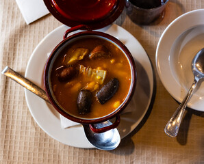 There is traditional Spanish dish on table - Fabada Asturian. Stewing soup cooking of beans, sausages and spices in clay pot