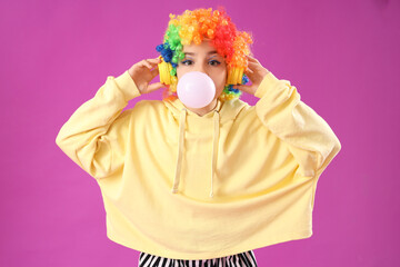 Trendy girl in clown costume blowing bubble gum while listening to music on purple background