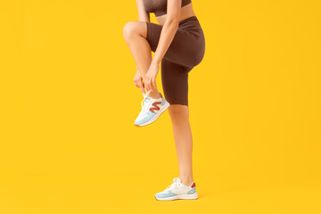 Young woman in cycling shorts tying shoe laces on yellow background - obrazy, fototapety, plakaty