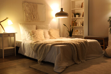 Interior of cozy bedroom with soft blankets on comfortable bed and glowing lamps at night