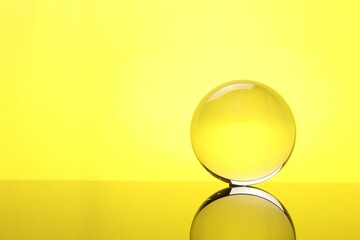 Transparent glass ball on mirror surface against yellow background. Space for text