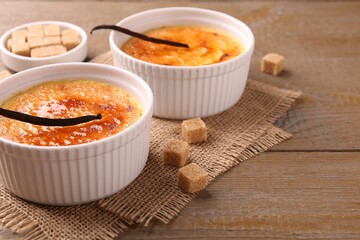 Delicious creme brulee in bowls, vanilla pods and sugar cubes on wooden table, closeup. Space for text