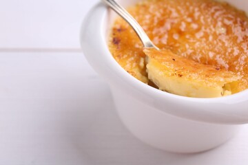 Taking delicious creme brulee with spoon from bowl at white wooden table, closeup. Space for text