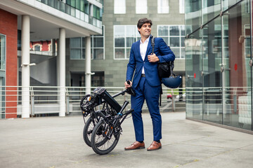 Businessman with backpack and bike helmet posing with folded electric folding bike in city...