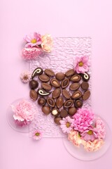 Heart made with delicious chocolate candies and beautiful flowers on pink background, flat lay