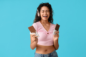 Beautiful young African-American woman in headphones with sweet chocolate bars on blue background - obrazy, fototapety, plakaty