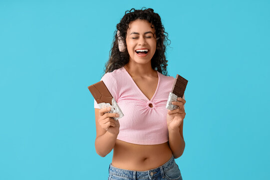 Beautiful young African-American woman in headphones with sweet chocolate bars on blue background
