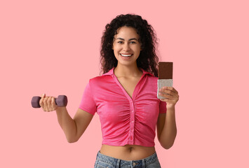 Happy young African-American woman with sweet chocolate bar and dumbbell on pink background - obrazy, fototapety, plakaty