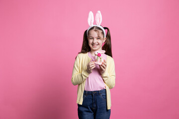 Obraz na płótnie Canvas Cute young kid posing with a pink stuffed rabbit toy in studio, presenting her decorations in time for easter celebration. Small girl with pigtails holding a colorful bunny on camera.