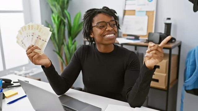 Joyous young black woman with dreadlocks in office, proudly brandishing danish krone banknotes. with a beaming smile and confident gesture, she points to one side.